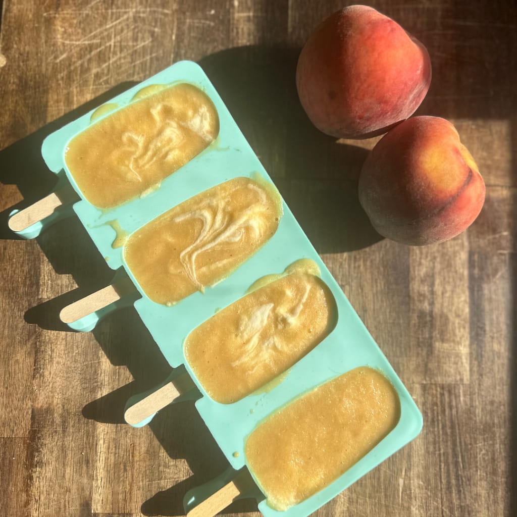 peaches and cream popsicles in a silicone mold with two fresh peaches on the counter