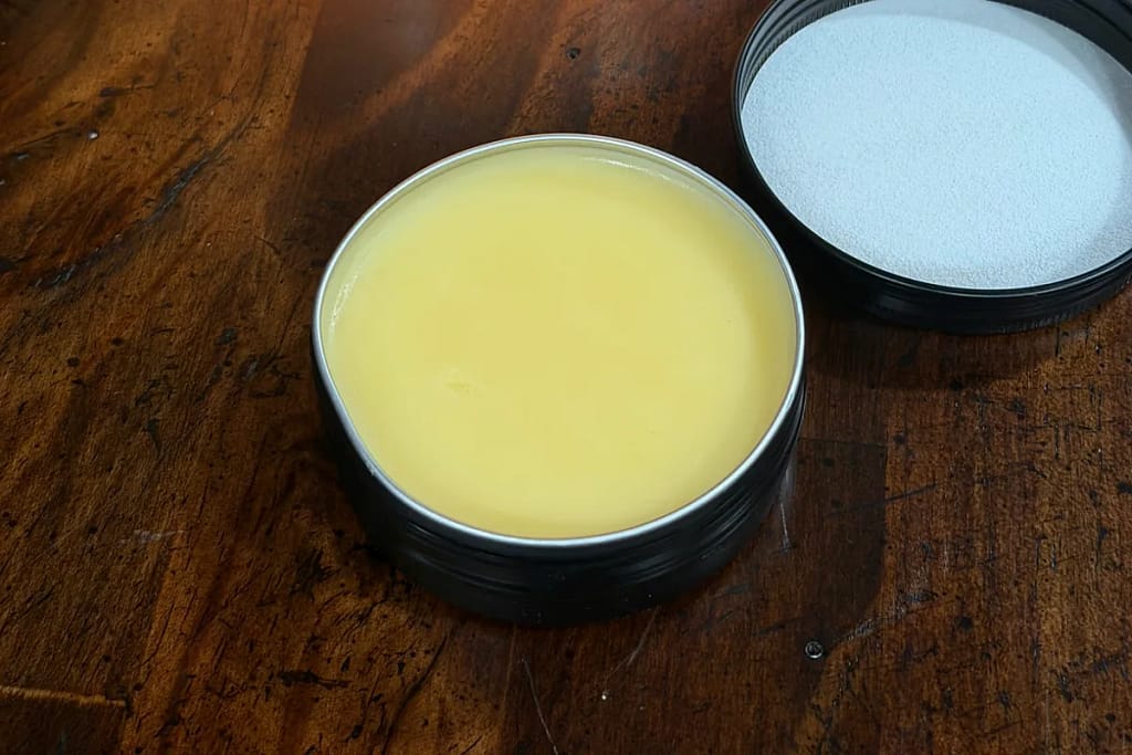 Open tin of pale yellow hand salve beside its lid on a wooden tabletop.