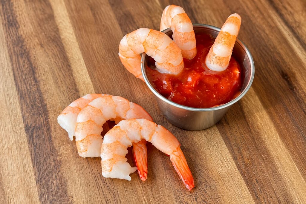 Shrimp served with homemade cocktail sauce in a small metal cup on a wooden board.