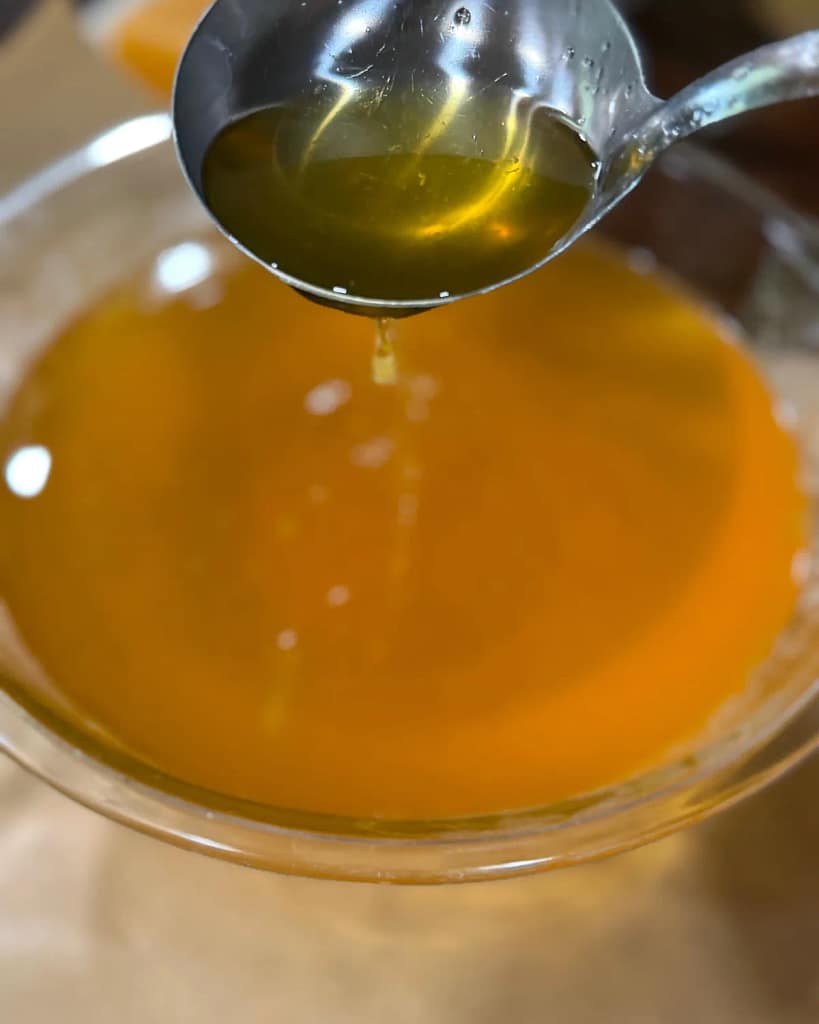 Ladle holding melted golden hand salve above a mixing bowl as a small drip falls back into the bowl.