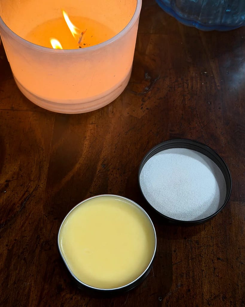 Open tin of pale yellow hand salve beside its lid on a wooden table, with a lit candle behind them.