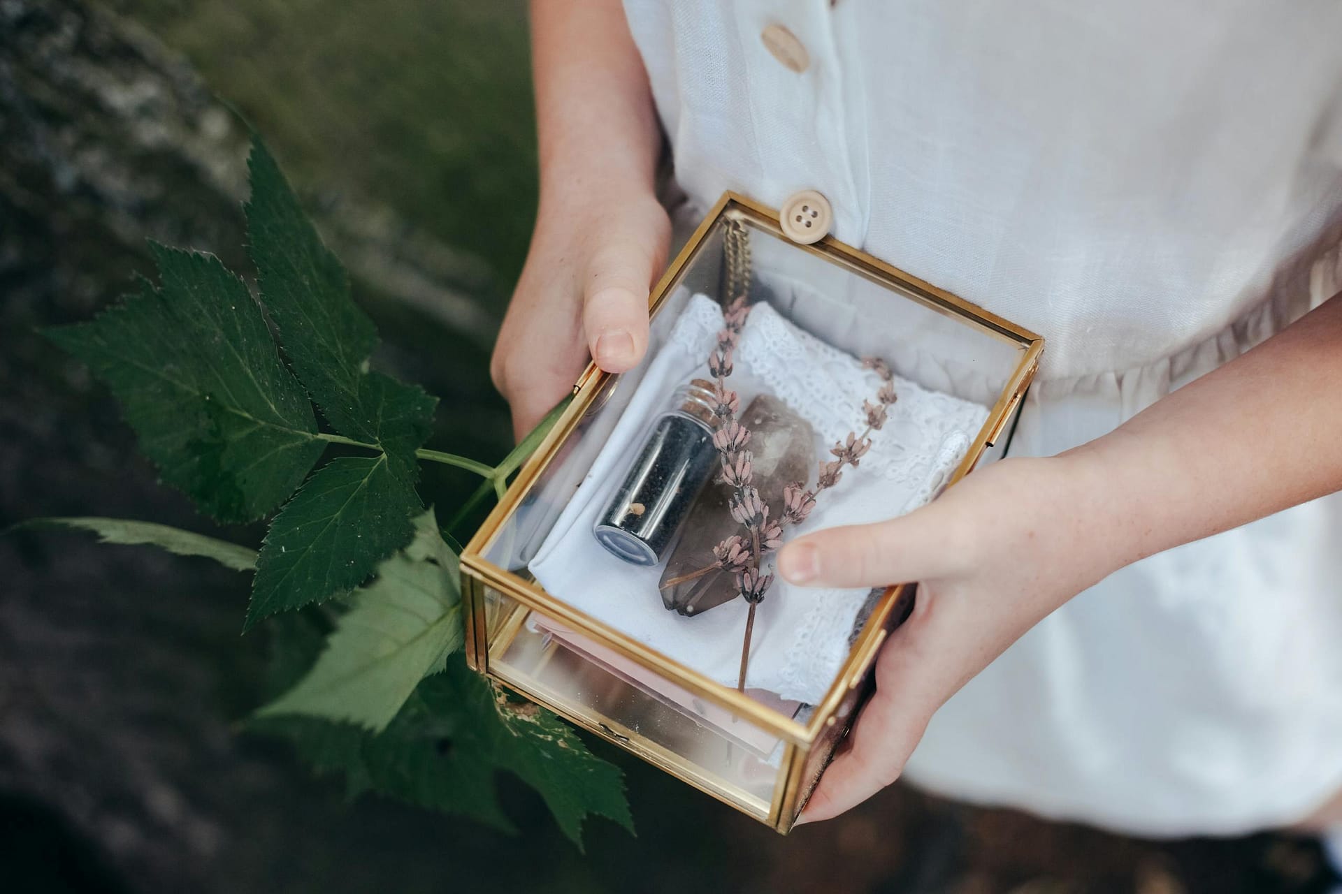 A young girl holds a decorative glass box with a vial and foliage, surrounded by nature.